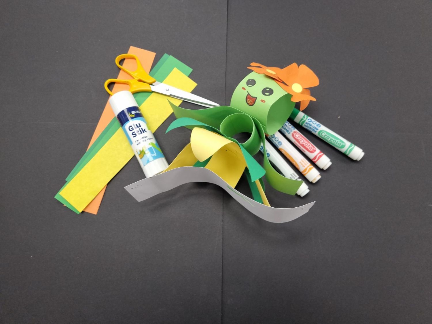 A green 3D paper character with a flower crown sits on a black surface next to scissors, a glue stick, colored paper strips, and Crayola markers.