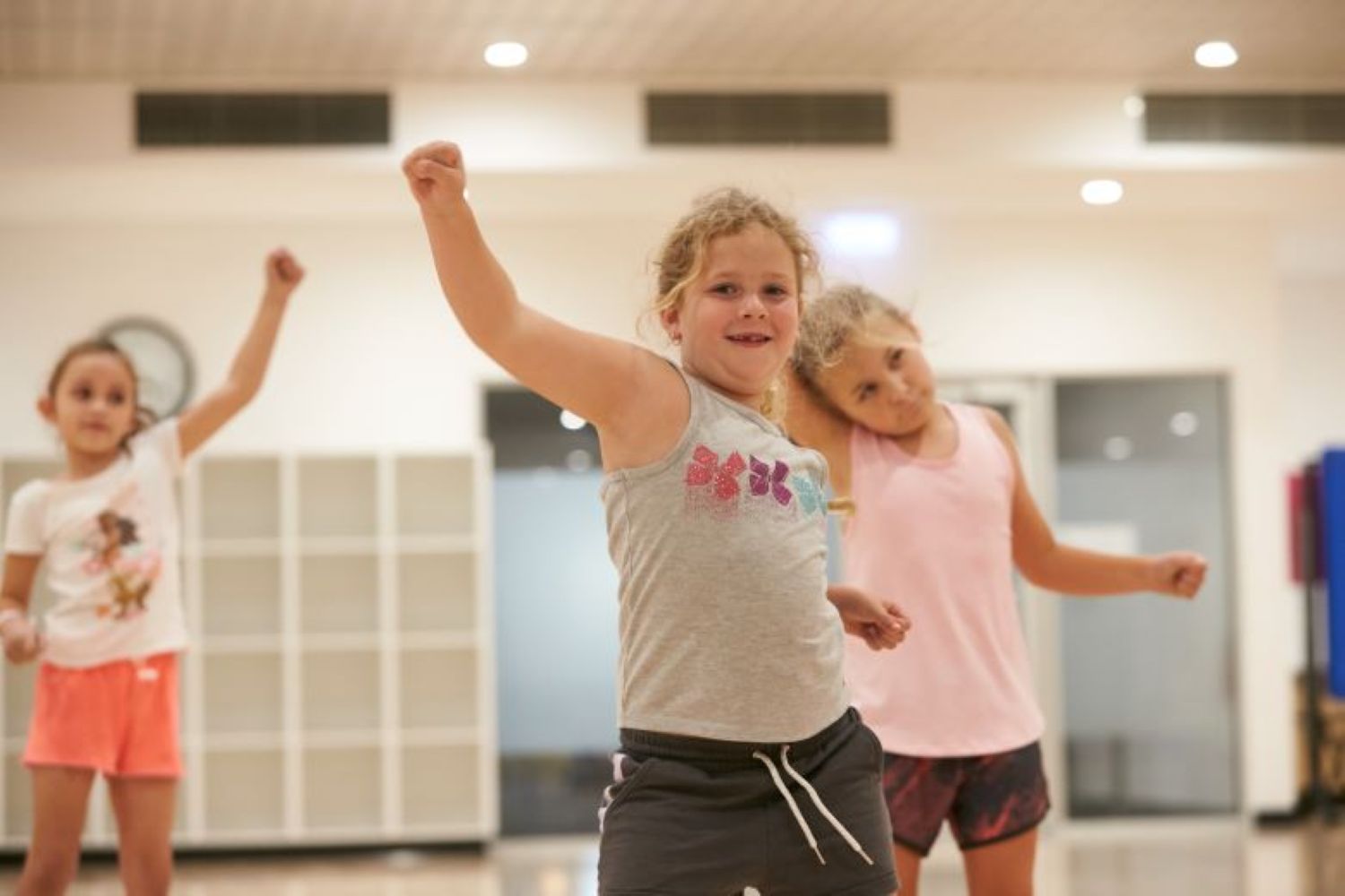 Three young girls practice choreography in a bright fitness studio. The girl in the center smiles with her arm raised in a fist, while others mimic the dance move behind her.