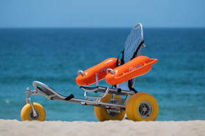 Beach wheelchair with bright orange flotation armrests and large yellow wheels on the sand with the ocean in the background.