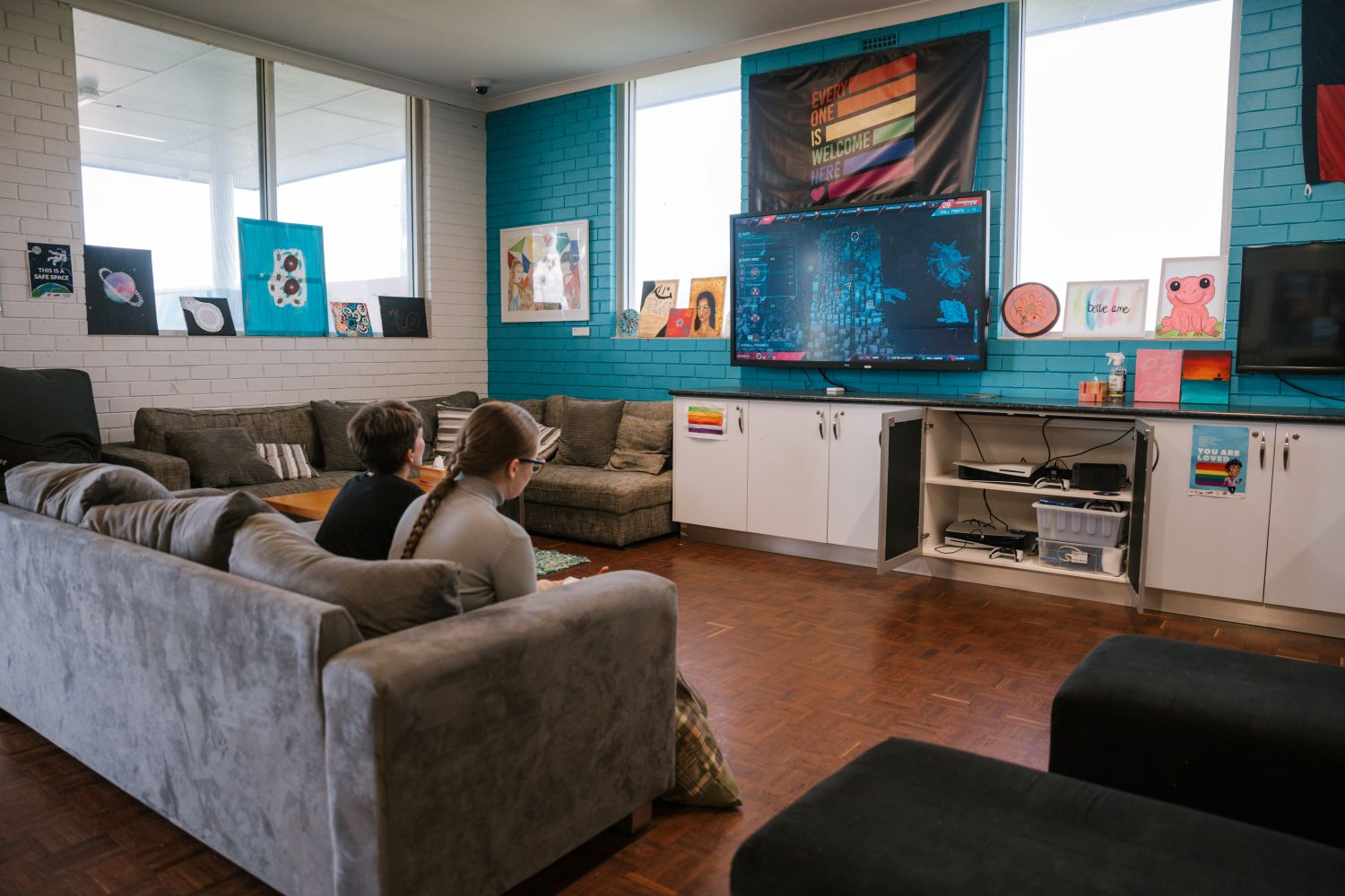 Two people sit on a couch watching a large wall‑mounted screen in a lounge room with artwork and storage cabinets.