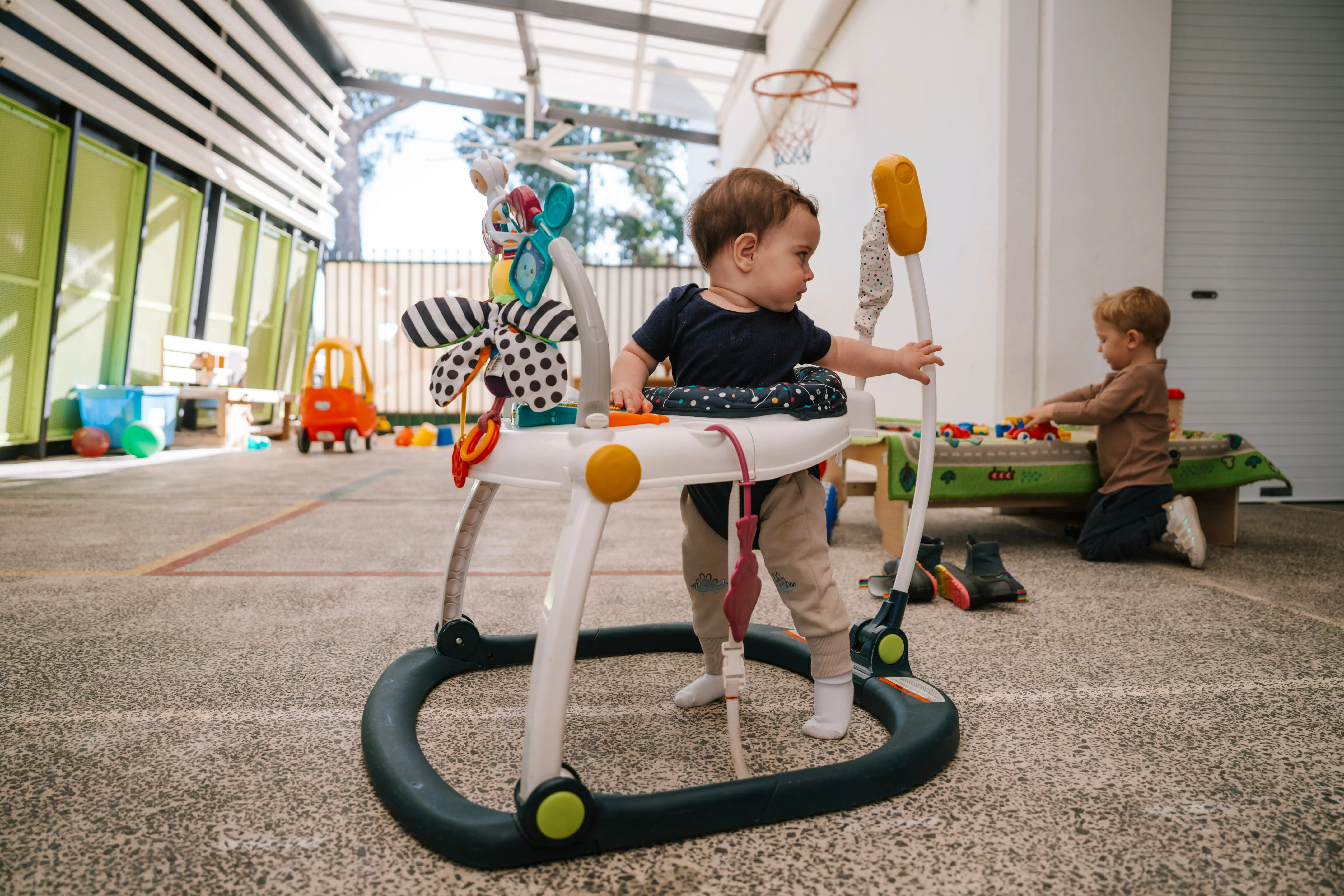 Baby standing in a play walker while another child plays nearby at the MARC crèche