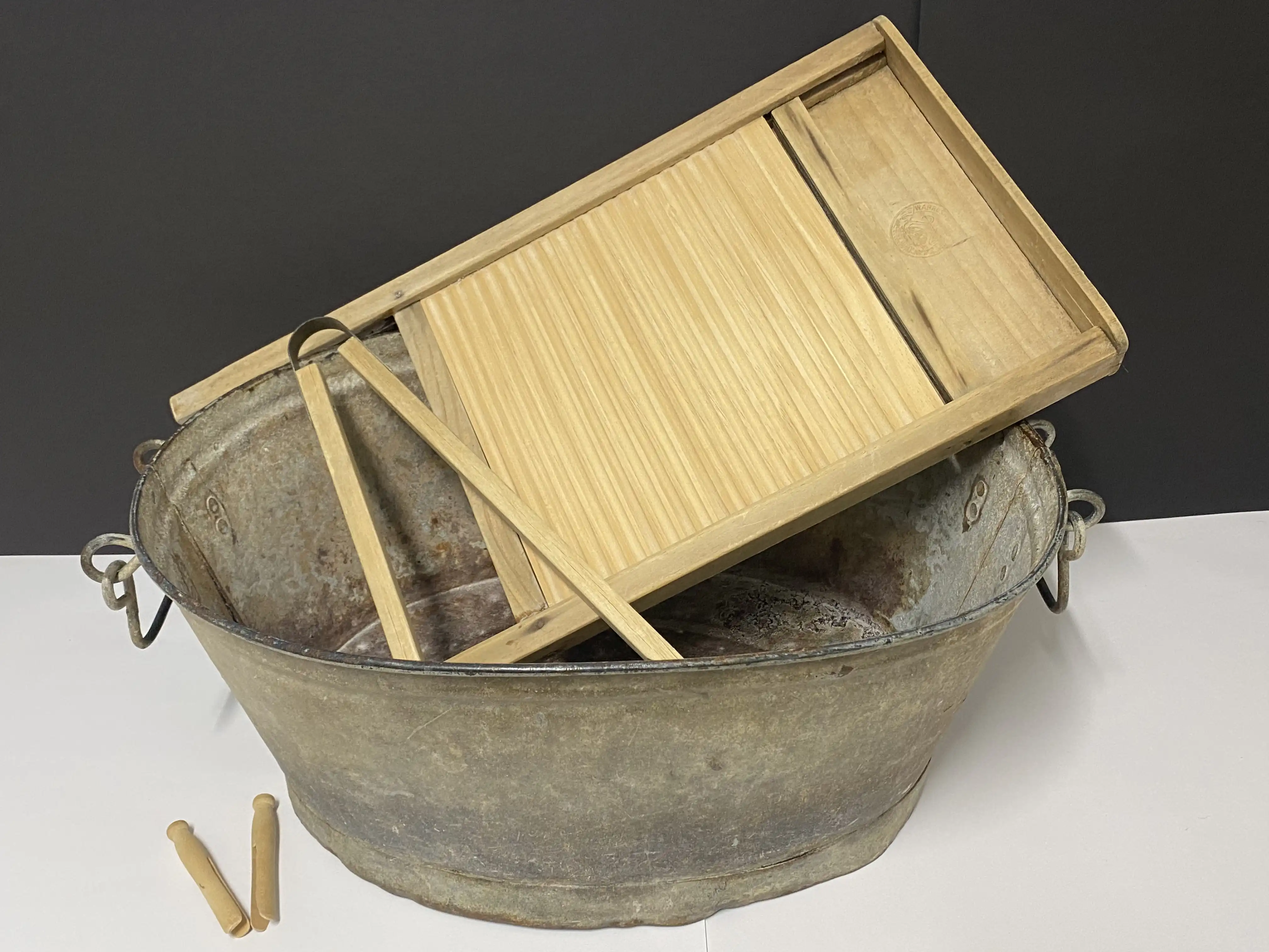 A large metal wash tub holding a wooden washboard and wooden laundry tools, with two wooden pegs placed beside it.