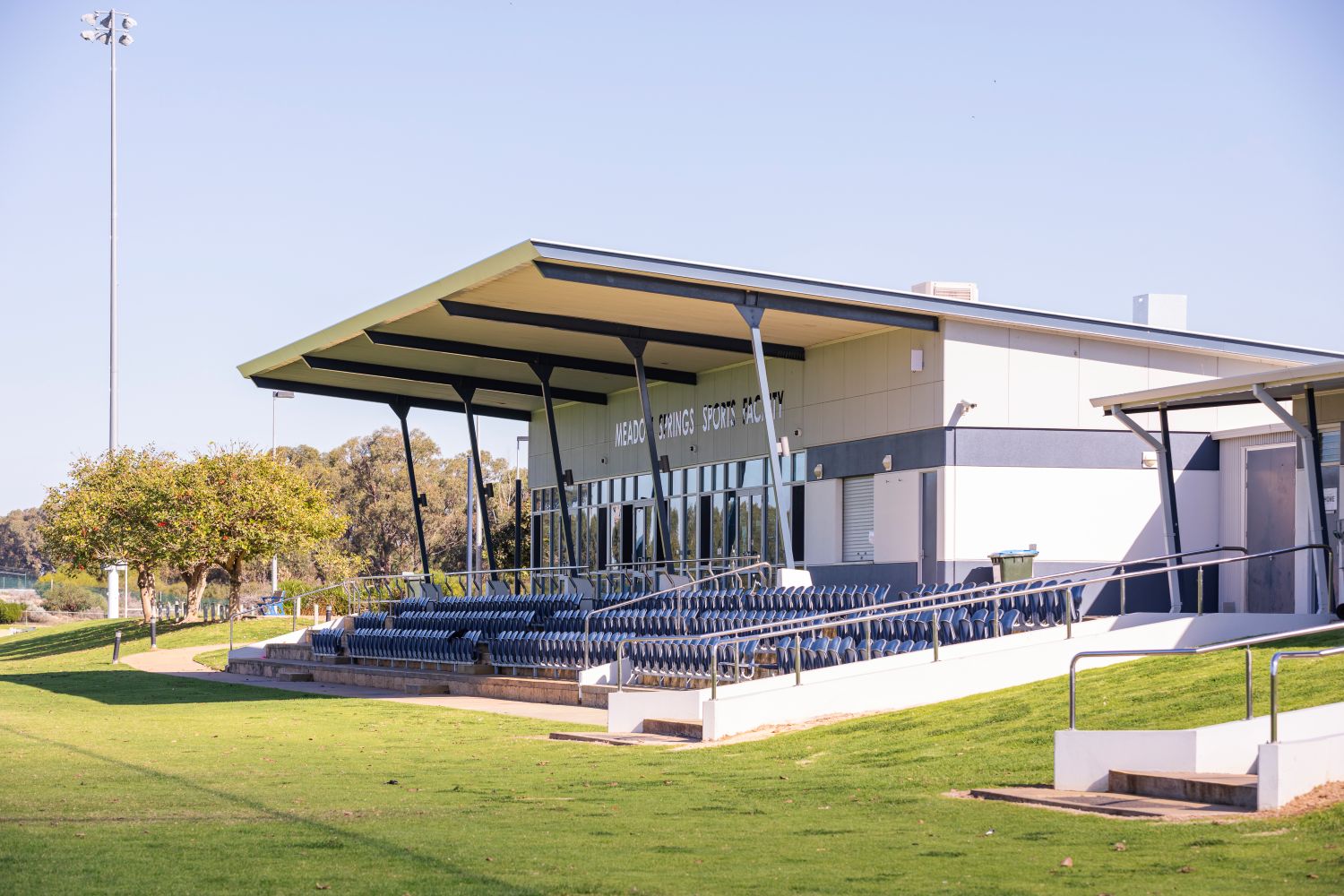 Meadow Springs Sports Facility exterior with covered spectator seating, tiered blue seats, grassed surrounds and the main building behind.